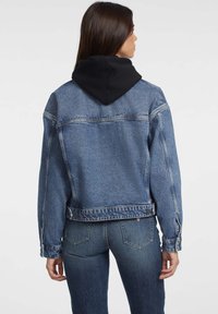 Jeans jakna z modrim pranjem, s črno kapuco. Kvadratna silhueta z vidnimi šivi in gumbom na dnu.