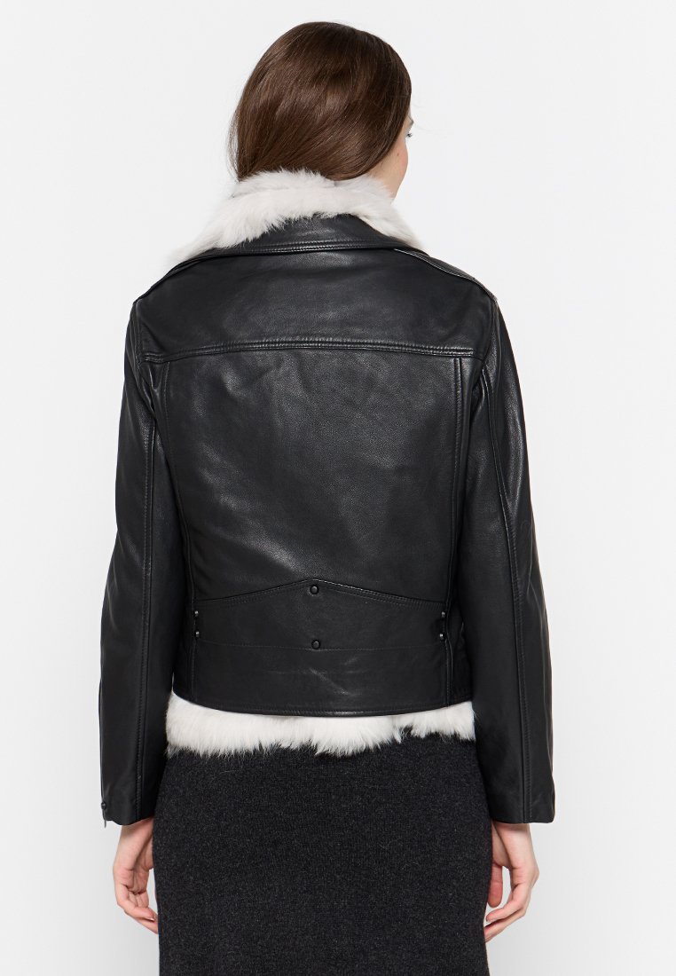 Frau trägt eine schwarze Lederjacke mit weißem Pelzkragen und -saum, steht mit dem Rücken zu einer schlichten weißen Hintergrund.