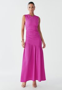 Vestido maxi fucsia con cuerpo ajustado y falda de volantes. Presenta un tejido texturizado y un diseño de escote en V. Combinado con zapatos de tacón de tiras.