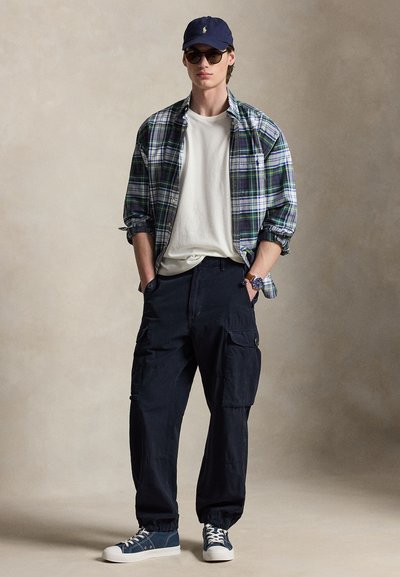 Jonge man met een blauwe baseballpet, donkere zonnebril, groen geruit overhemd over een wit T-shirt, zwarte cargobroek en blauwe sneakers, staand met handen in de zakken.