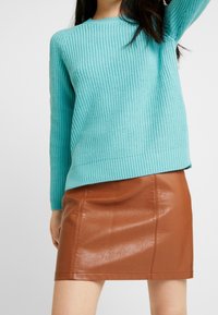 Pull en tricot turquoise avec texture côtelée et manches longues, associé à une mini-jupe en simili cuir marron à finition lisse et coutures apparentes.
