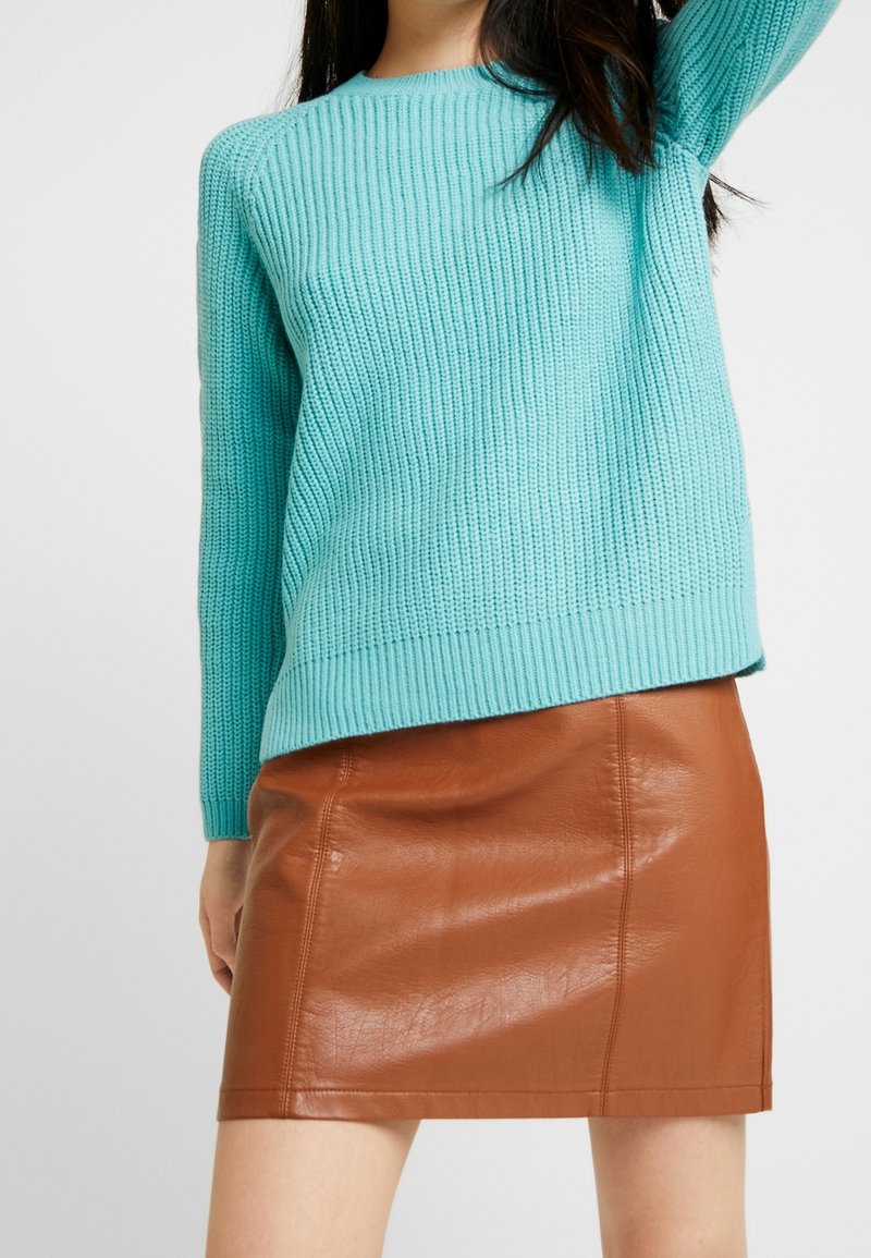 Pull en tricot turquoise avec texture côtelée et manches longues, associé à une mini-jupe en simili cuir marron à finition lisse et coutures apparentes.