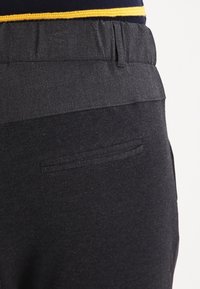 Donkergrijze broek met een vlakke tailleband, elastische bovenkant en een klein achterzakje. De stof lijkt glad met een subtiele textuur.
