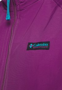 Veste violette avec fermeture éclair violette et cordons turquoise, arborant un patch logo noir Columbia Sportswear Company sur la poitrine.