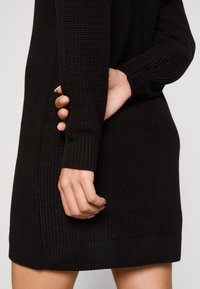 Robe-pull noire tricotée avec une surface texturée, des poignets côtelés et une finition ourlet. Main placée sur le côté près de la couture.
