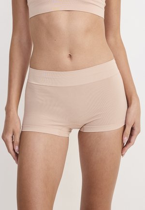 Frau trägt hautfarbene nahtlose Boyshorts und passendes kurzes Oberteil, steht vor einfachem hellem Hintergrund.