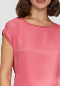 Roze blouse met korte mouwen en een ronde halslijn. De stof lijkt glad en zacht, met een subtiele glans en eenvoudige ontwerpelementen.