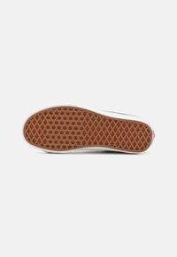 Zapatilla blanca con una suela de goma texturizada, que presenta un patrón enrejado y una suela marrón. Detalle en rojo en el talón.