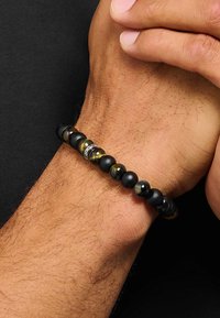 Schwarzes Perlenarmband mit runden matten und polierten Steinen, durchsetzt mit metallischen Akzenten; getragen an einem Handgelenk.