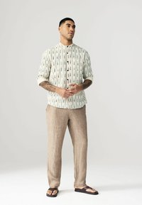 Chemise en coton à boutons avec une base blanc cassé, ornée de motifs de flèches noires. Associée à un pantalon en lin beige et des tongs marron.