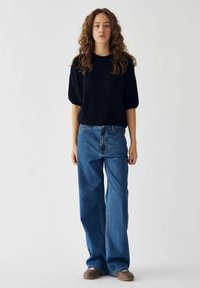 Fekete kötött pulóver rövid, fodros ujjal, kék, bő szárú farmerrel párosítva. Laza stílus, puha anyag, kényelmes szabás. Barna cipő.