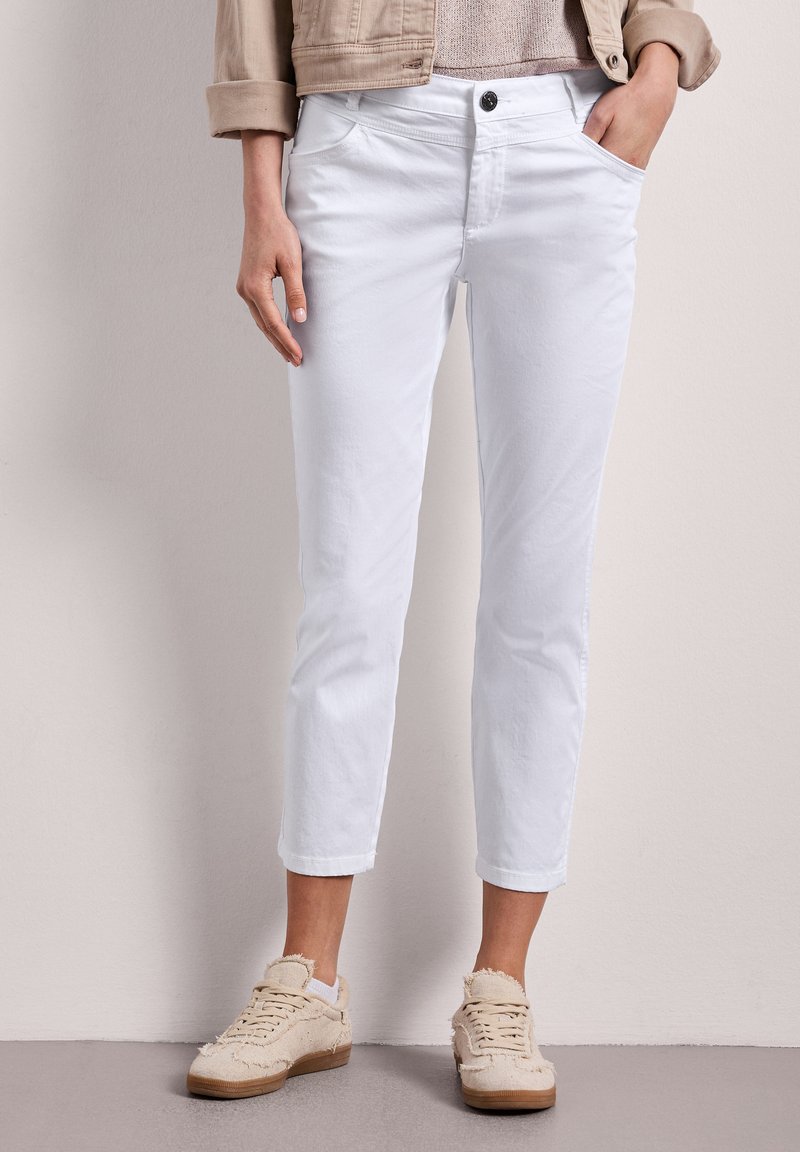 Femme portant un pantalon blanc court, des baskets beiges texturées et une veste beige sur un fond clair uni.