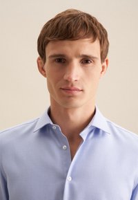 Camicia azzurra chiara con un motivo tessuto, dotata di colletto aperto e piccoli bottoni bianchi, indossata da un uomo con capelli corti.