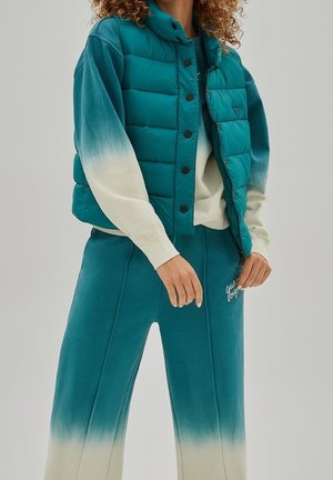 Person trägt eine türkisfarbene Steppweste über einem türkis-weißen Sweatshirt mit Farbverlauf und passende Sweatpants mit Farbverlauf sowie weißem gesticktem Logo.