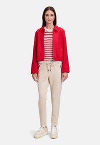 Rote, aus Wolle gemischte, kurz geschnittene Jacke, darunter ein rot-weiß gestreifter Pullover, beige Sweatpants mit Kordelzug und weiße Sneaker mit beigen Akzenten.