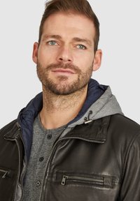 Chaqueta de cuero negra con capucha gris, bolsillos con cremallera, herrajes plateados, acabado texturizado y camiseta henley gris debajo.