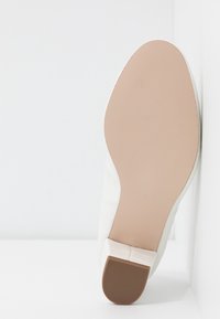 Semelle d'une chaussure blanche avec une bande de roulement beige lisse et un petit talon carré marron, vue de dessous sur un fond blanc.