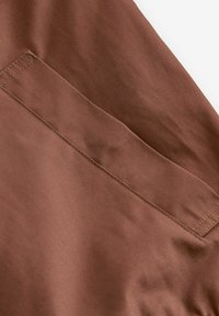 Tela marrón con una textura suave, que presenta un bolsillo visible y una costura ordenada. El diseño tiene un brillo sutil y líneas diagonales.