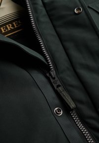 Ciemnozielona kurtka z widocznym czarnym zamkiem błyskawicznym, teksturowanym materiałem i lekko błyszczącym wykończeniem. Zauważalny metalowy zatrzask.