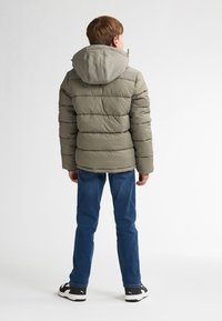 Giubbotto a vento verde tenue con cuciture orizzontali, cappuccio e cerniera. Abbinato a jeans blu e scarpe da ginnastica nere.