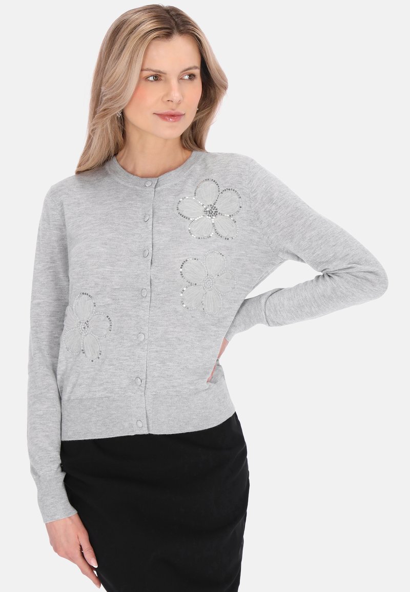 Blondynka w jasnoszarym, zapinanym na guziki kardiganie z kwiatowymi wzorami z cekinami, w czarnej spódnicy, ręka na biodrze, spogląda w lewo.