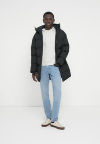 Μαύρο μπουφάν τύπου puffer με κουκούλα, φορεμένο πάνω από ανοιχτό γκρι φούτερ, σε συνδυασμό με ανοιχτό μπλε τζιν και μπεζ αθλητικά παπούτσια. Απλός, μοντέρνος σχεδιασμός.