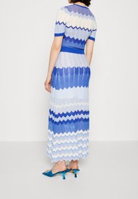 Maxi robe à motifs zigzag bleu et blanc avec manches courtes, tissu en maille texturée, et une taille cintrée. Associée à des mules à talons bleues.