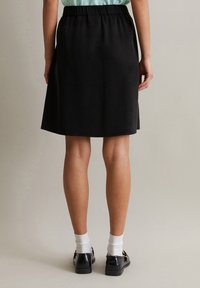 Schwarzer knielanger Rock mit elastischem Bund, glatter Textur und schlichter Gestaltung. Getragen mit schwarzen Schuhen und weißen Socken.