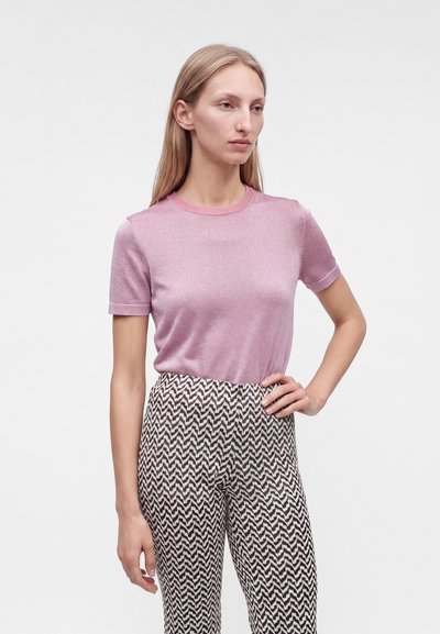 Femme aux longs cheveux blonds porte un haut rose clair à manches courtes et un pantalon à motif zigzag noir et blanc, debout main sur la hanche.