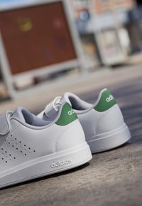 Baskets blanches avec un dessus en cuir lisse, accent vert au niveau du talon, design perforé pour la respirabilité et semelle en caoutchouc texturée.