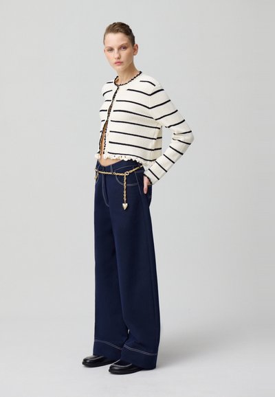 Cardigan court rayé blanc et marine, texture tricotée, avec un ourlet festonné. Assorti à un pantalon large marine avec une ceinture en chaîne dorée.