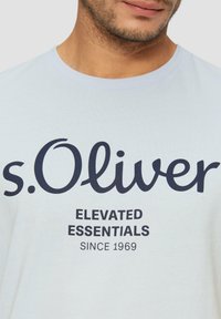 Hele sinine puuvillane T-särk tumesinise trüki ning brändi nime "s.Oliver" ja tekstiga "ELEVATED ESSENTIALS" rasvases fondis. Pehme tekstuur.