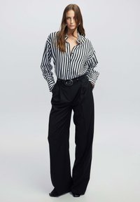 Gestreifte Bluse mit schwarz-weißen vertikalen Streifen, schwarze hochtaillierte Hose mit weitem Bein und einem eleganten schwarzen Gürtel mit runder Schnalle.