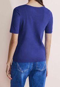 Lila t-shirt med kort ärm och rund halsringning, tillverkad av slätt bomullstyg, i kombination med blå denimbyxor med bakfickor.