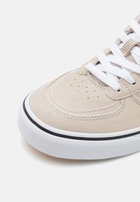Beige suede sneaker with white laces and a textured sole, featuring a contrasting black stripe along the edge and perforations on the toe.