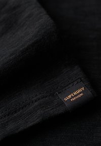 Zwart stof met een fijne textuur, voorzien van een zichtbaar "Superdry Premium" label in koperkleurige stiksel. Focus op materiaal- en ontwerpdétails.