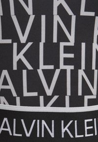 Sort undertøjsbånd med fremtrædende hvide bogstaver "CALVIN KLEIN" i forskellige størrelser, med kontrasterende grå stribeaccenter.