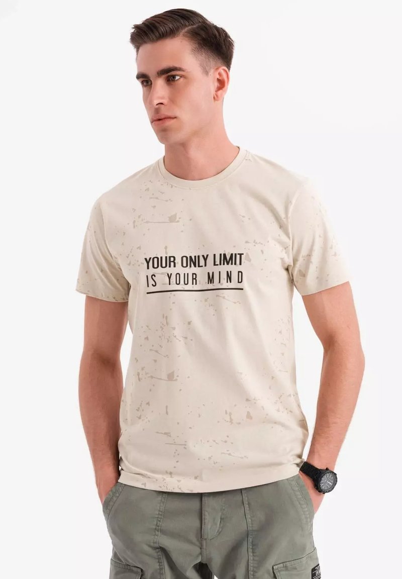 Beige t-shirt med en spraglet tekstur. Udstyret med sort tekst, der siger "DIN ENESTE GRÆNSE ER DIN BEVIDSTHED." Korte ærmer, rund halsudskæring.