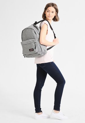 Mochila gris con una tela texturizada, que cuenta con un bolsillo frontal y correas ajustables, llevada por una persona con atuendo casual.