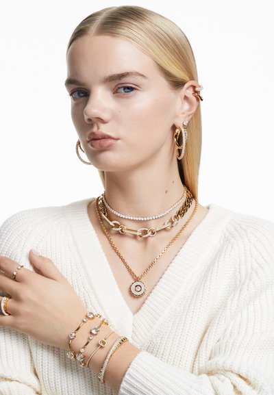 Bijoux en or comprenant des colliers superposés, des boucles d'oreilles créoles et plusieurs bracelets. Or texturé et pierres transparentes, sur un pull blanc.