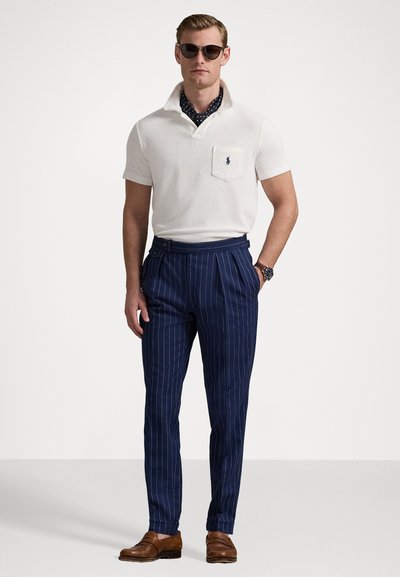Polo shirt blanc avec une poche ; pantalon rayé bleu et blanc ; mocassins marron ; lunettes de soleil foncées ; écharpe à pois ; coupe élégante et ajustée.