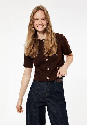 Giovane donna con lunghi capelli ondulati che sorride, indossa un top marrone a maniche corte con bottoni e pantaloni scuri a vita alta, in piedi su sfondo bianco.