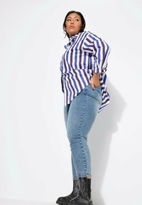 Camisa de botones a rayas moradas y blancas, vaqueros ajustados de color azul claro y botas de tobillo negras con costuras decorativas, posando sobre un fondo blanco.
