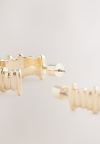 Gold-toned earrings with a ribbed texture and rounded ends, featuring a push-back fastening. Smooth, shiny finish with no visible embellishments.