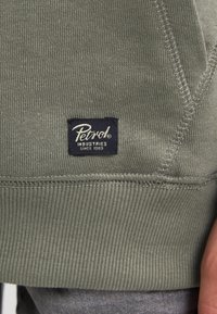 Tejido de sudadera acanalado verde con una etiqueta azul marino que muestra "Petrol Industries Since 1989", con costuras texturizadas y puños sin costuras.