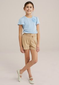 Ljusblå ribbad kortärmad skjorta med blommor, tillsammans med beiga högmidjade veckade shorts och ljusgröna skor med detaljer.