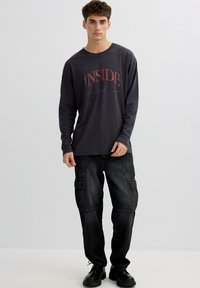 Joven de pie, vestido con una camisa de manga larga oscura con el texto "INSIDE" y pantalones cargo negros con zapatos negros de cordones, sobre un fondo gris claro.