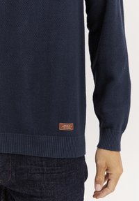 Pull en tricot bleu marine avec un motif texturé, des poignets et un ourlet côtelés. Il présente une petite étiquette en cuir marron sur le côté inférieur.