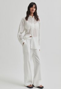 Bela, perforirana srajca s dolgimi rokavi, uparjena z ohlapnimi hlačami enake barve. Ravne sandale dopolnjujejo oblačilo.