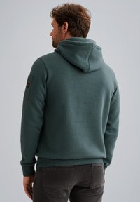 Kapuzensweatshirt aus gedämpftem Teal-Stoff, mit strukturiertem Finish, gerippten Bündchen und einem Patch am linken Ärmels. Rückansicht.